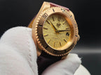 A close-up of The Midas SJT Original wristwatch with a textured gold dial, black bezel markings, and a dark brown leather strap, held by a white gloved hand against a dark background.