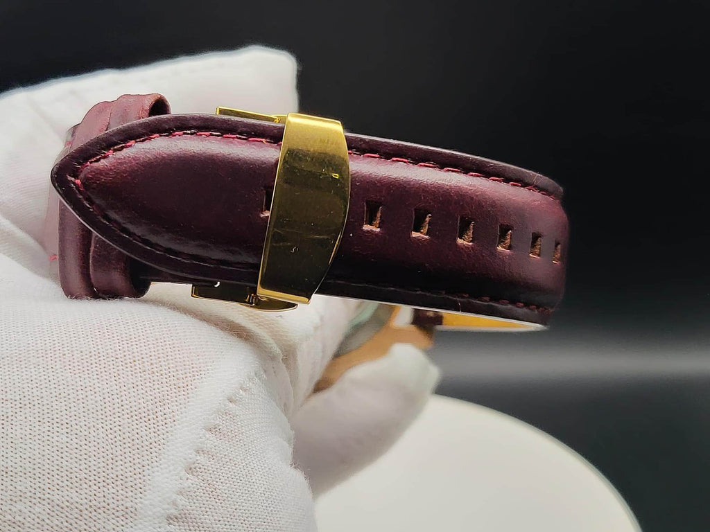 A close-up of a dark burgundy leather watch strap with gold-toned hardware, for The Midas SJT Original wristwatch, held by a white gloved hand against a dark background.