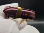 A close-up of a dark burgundy leather watch strap with gold-toned hardware, for The Midas SJT Original wristwatch, held by a white gloved hand against a dark background.
