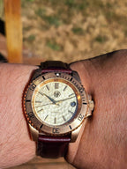 A close-up of The Midas SJT Original wristwatch with a bronze-toned bezel, cream-colored textured dial, and a dark burgundy leather strap, shown worn on a person's wrist against a blurred outdoor background.