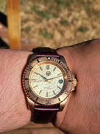 The Midas SJT Original wristwatch with a bronze-toned case, cream-colored dial, and dark brown leather strap, shown worn on a person's wrist against an outdoor blurred background.