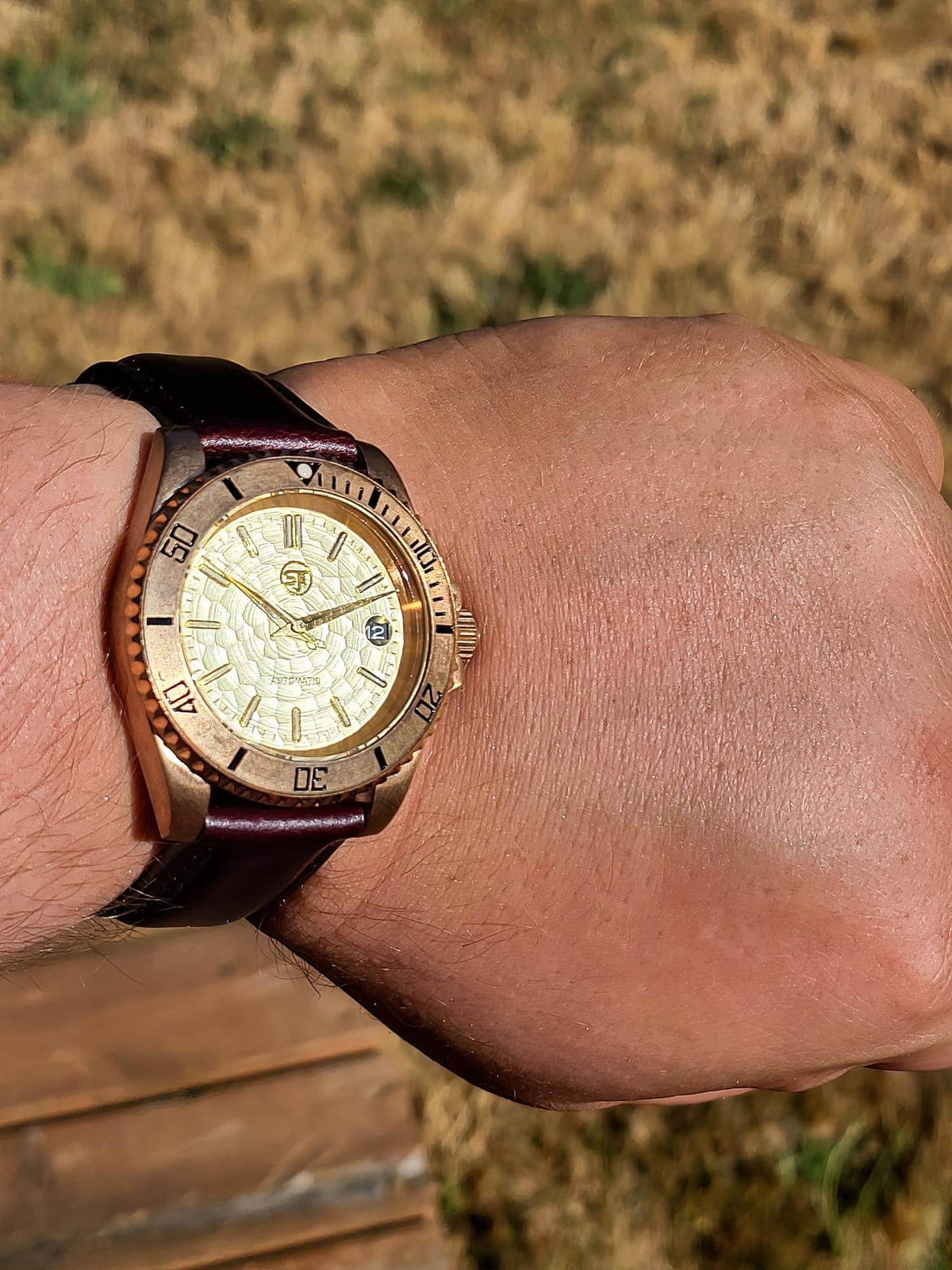 A close-up of The Midas SJT Original wristwatch with a rose gold-toned case, textured cream dial, and dark brown leather strap, shown worn on a person's wrist against an outdoor background.