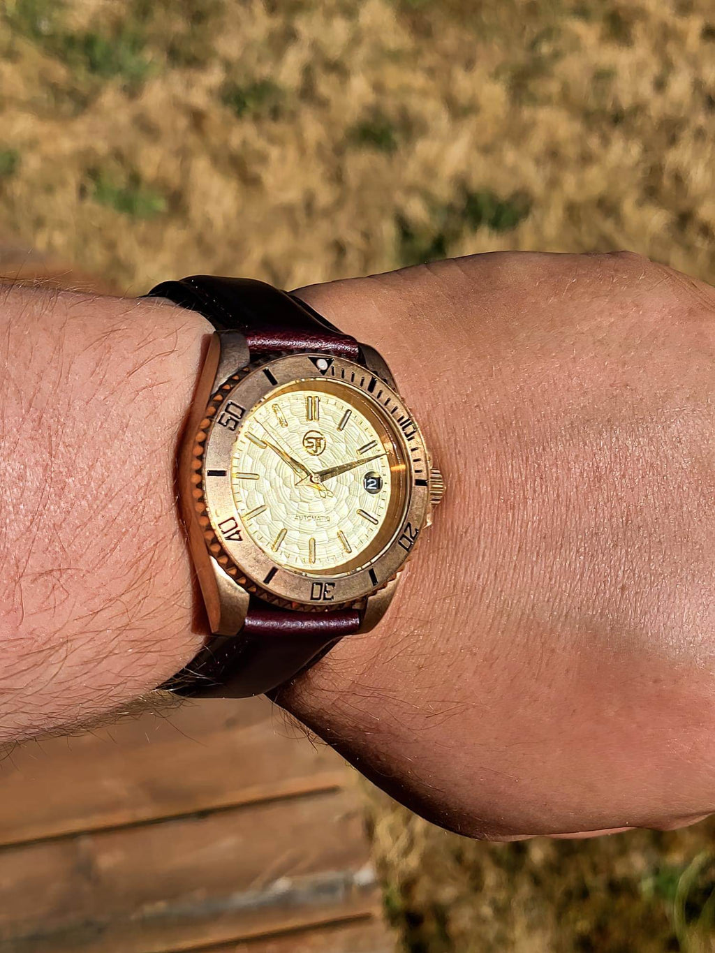 A close-up of The Midas SJT Original wristwatch with a gold-toned textured dial, a gold bezel with black markings, and a dark brown leather strap, worn on a person's wrist against an outdoor background.