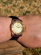 A close-up of The Midas SJT Original wristwatch with a gold-toned textured dial, a gold bezel with black markings, and a dark brown leather strap, worn on a person's wrist against an outdoor background.