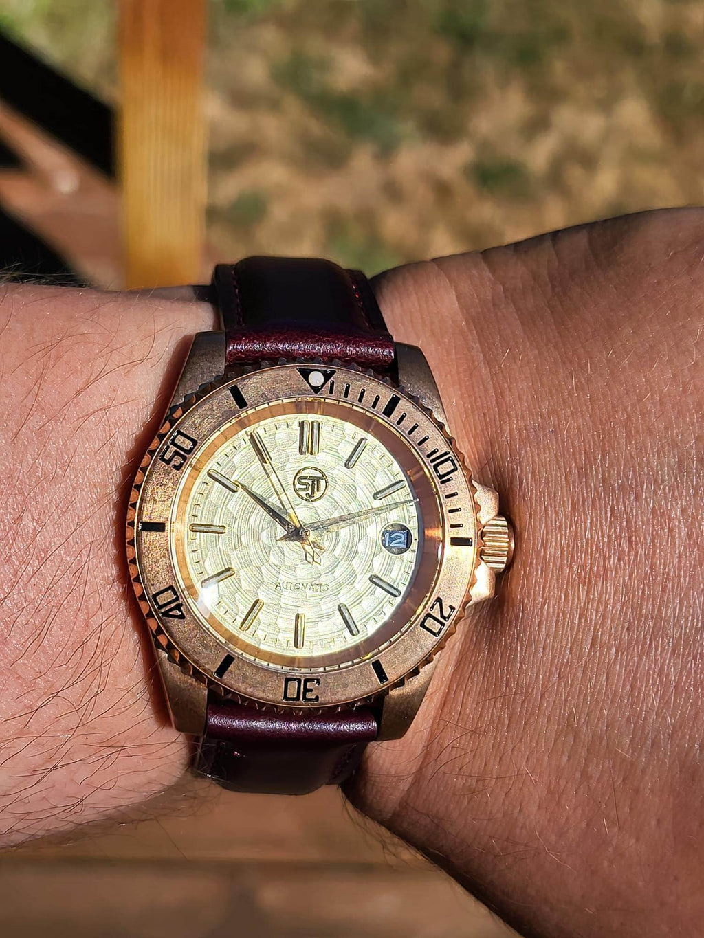 The Midas SJT Original wristwatch with a bronze-toned case, textured cream dial, and dark burgundy leather strap, shown worn on a person's wrist against a blurred outdoor background.