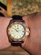 The Midas SJT Original wristwatch with a bronze-toned case, textured cream dial, and dark burgundy leather strap, shown worn on a person's wrist against a blurred outdoor background.