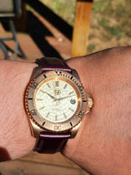 The Midas SJT Original wristwatch with a gold-toned case, cream dial, and dark burgundy leather strap worn on a person's wrist in an outdoor setting.