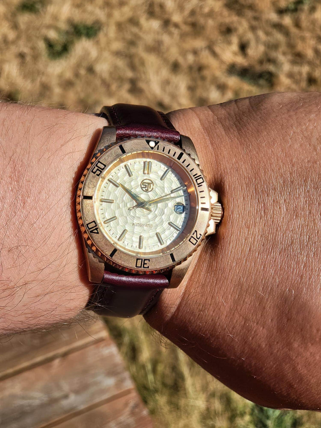 A close-up of a wrist wearing The Midas SJT Original wristwatch outdoors. The watch has a rose gold-toned case, a textured white dial with gold-tone markers, and a dark brown leather strap.