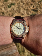 The Midas SJT Original wristwatch with a textured cream dial, bronze-toned bezel and case, and a dark burgundy leather strap, shown worn on a person's wrist against a blurred outdoor background.