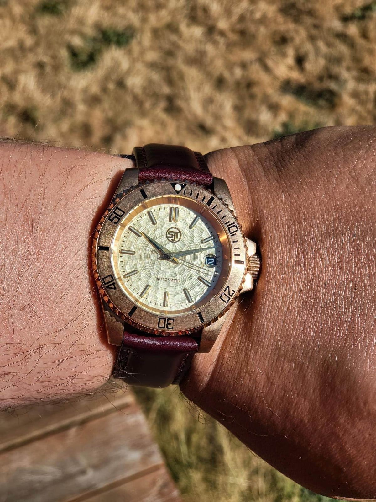 The Midas SJT Original wristwatch with a textured cream dial, bronze-toned bezel and case, and a dark burgundy leather strap, shown worn on a person's wrist against a blurred outdoor background.