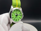 A close-up of The P-A-R-T-Y? SJT Original wristwatch with a bright green dial and matching green fabric strap held by a white gloved hand against a dark studio background.