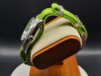 The P-A-R-T-Y? SJT Original wristwatch with a green woven strap, shown from the side resting on a wooden display stand with a cream cushion against a dark background.