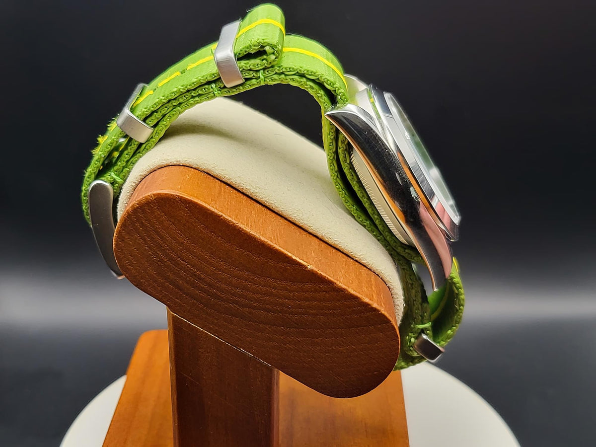 A close-up of The P-A-R-T-Y? SJT Original wristwatch with a bright green textured strap and silver-toned case, displayed on a wooden stand against a dark studio background.