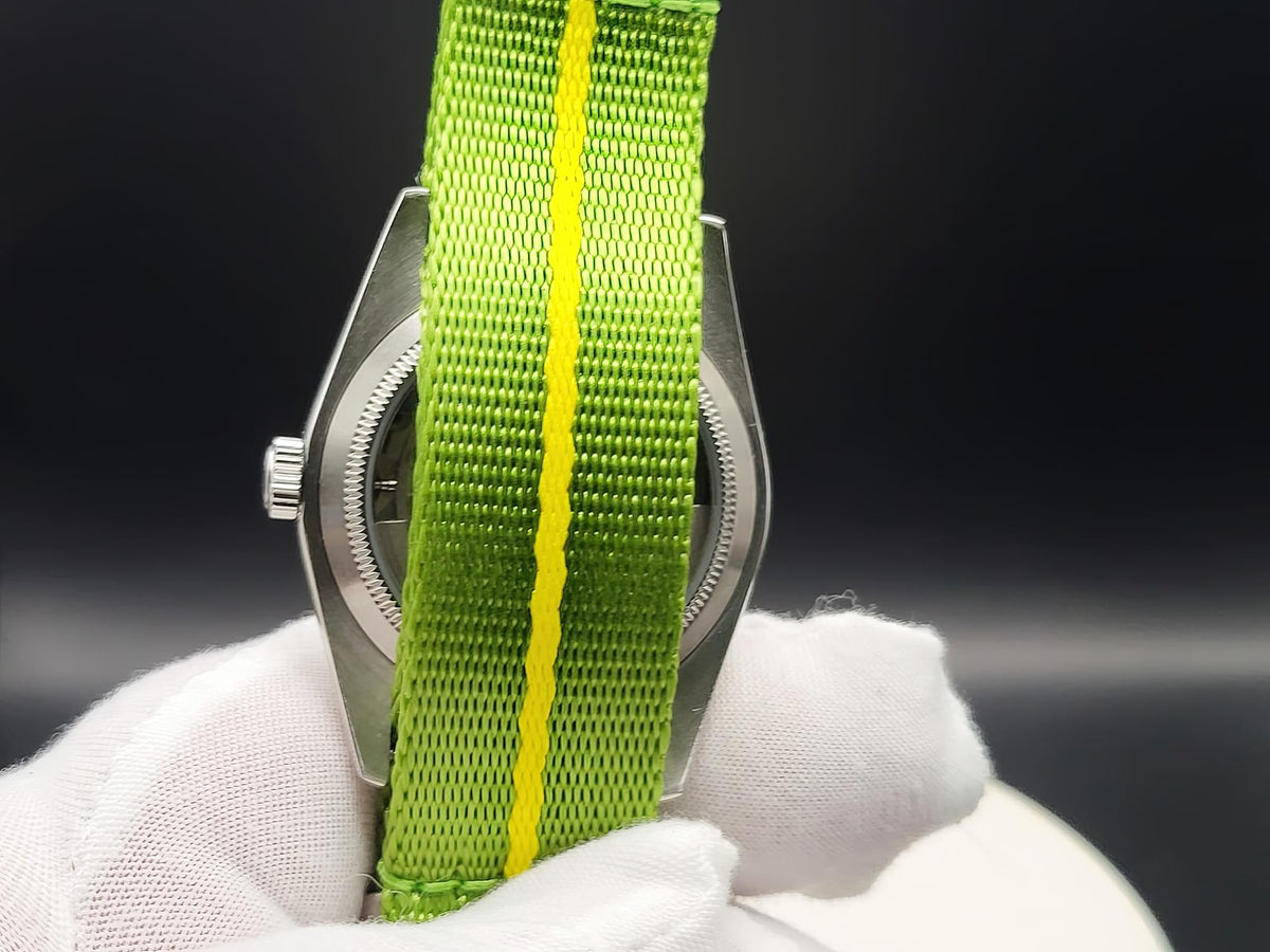 A close-up of a silver-toned watch case with a bright green woven strap featuring a single yellow stripe, for The P-A-R-T-Y? SJT Original wristwatch, held by a white gloved hand against a dark background.