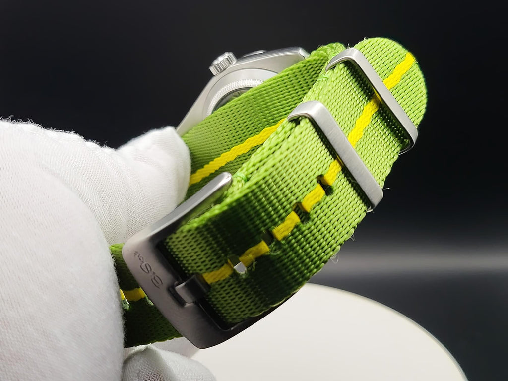 A close-up of a green nylon watch strap with yellow stitching and silver metal hardware, for The P-A-R-T-Y? SJT Original wristwatch, held by a white gloved hand against a dark background.