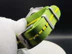 A close-up of a green nylon watch strap with yellow stitching and silver metal hardware, for The P-A-R-T-Y? SJT Original wristwatch, held by a white gloved hand against a dark background.