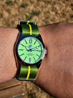 A close-up of a wrist wearing The P-A-R-T-Y? SJT Original wristwatch with a bright green dial and a green-and-yellow striped fabric strap, photographed outdoors against a blurred tan ground background.