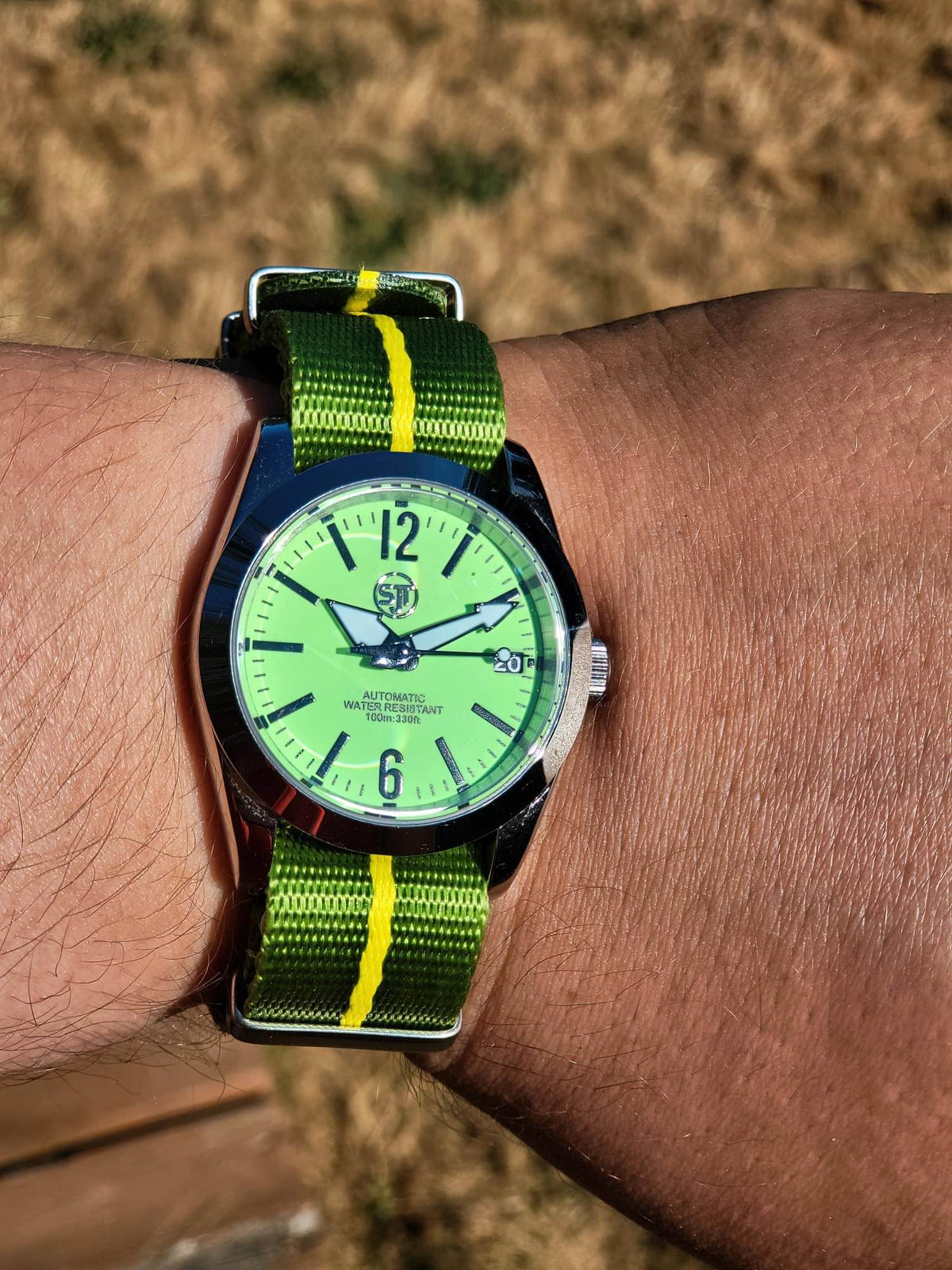 A close-up of a wrist wearing The P-A-R-T-Y? SJT Original wristwatch with a bright green dial and a green-and-yellow striped fabric strap, photographed outdoors against a blurred tan ground background.