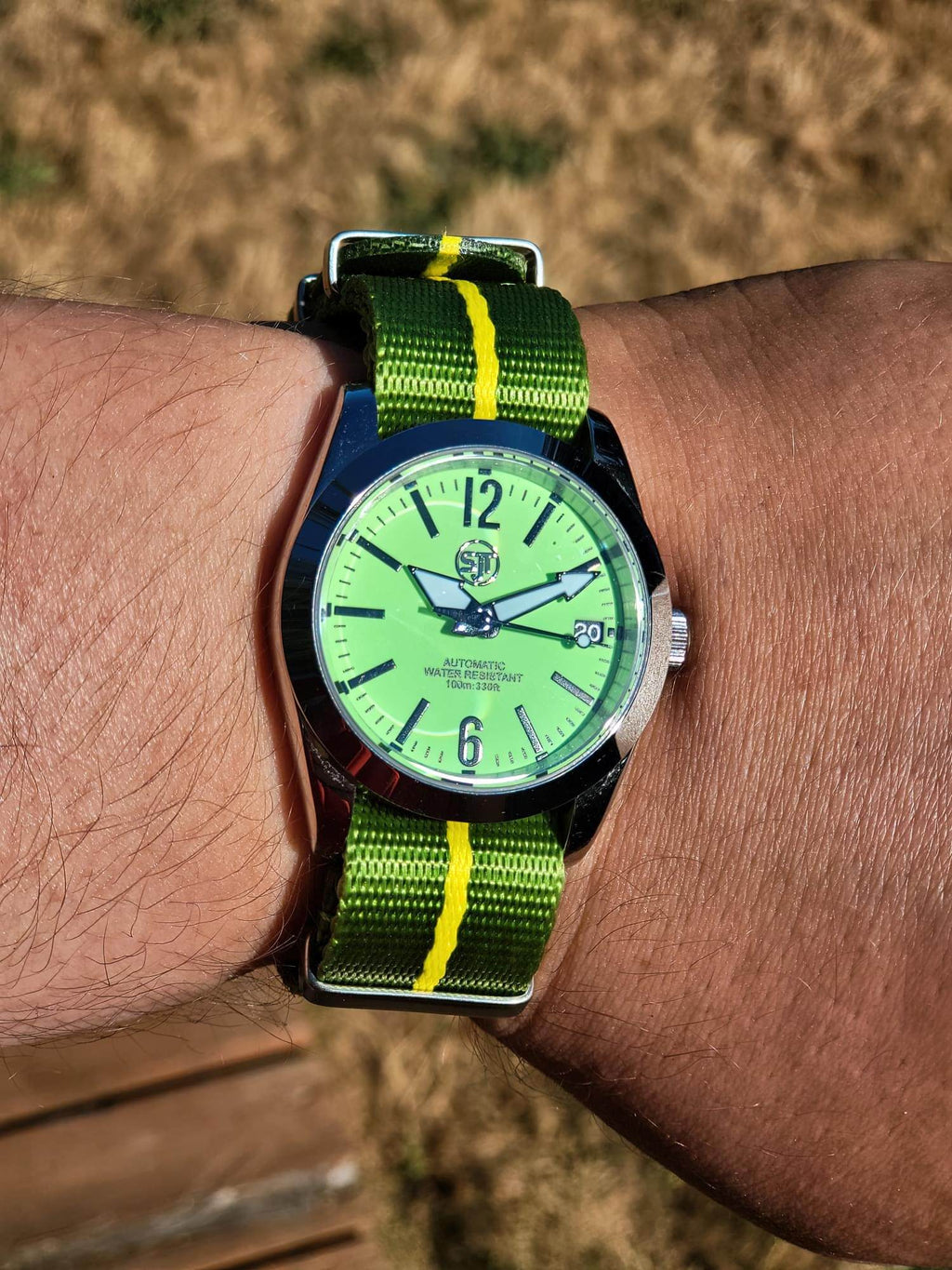 The P-A-R-T-Y? SJT Original wristwatch with a bright green dial and a green nylon strap with yellow stripes, worn on a person's wrist against a blurred outdoor background.