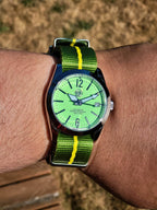 The P-A-R-T-Y? SJT Original wristwatch with a bright green dial and a green nylon strap with yellow stripes, worn on a person's wrist against a blurred outdoor background.