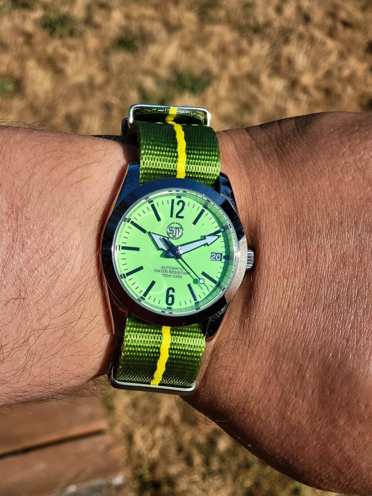 The P-A-R-T-Y? SJT Original wristwatch with a bright green dial and a green-and-yellow striped nylon strap, worn on a person's wrist against an outdoor dry grass background.