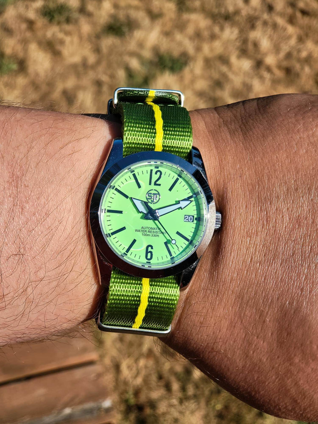 The P-A-R-T-Y? SJT Original wristwatch with a bright green dial and a green-and-yellow striped fabric strap, worn on a person's wrist against a blurred outdoor background.
