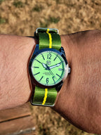 The P-A-R-T-Y? SJT Original wristwatch with a bright green dial and a green-and-yellow striped fabric strap, worn on a person's wrist against a blurred outdoor background.