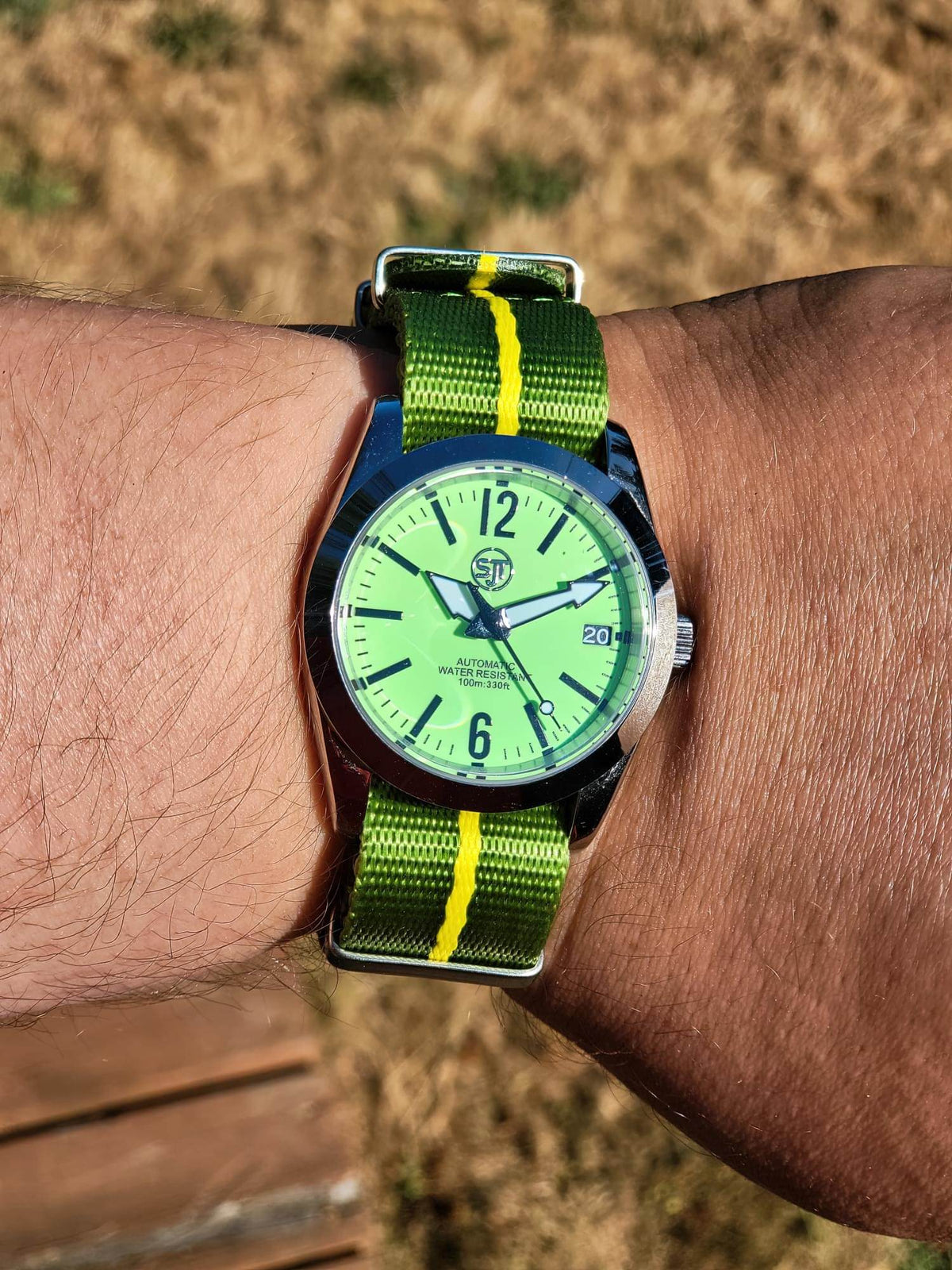 The P-A-R-T-Y? SJT Original wristwatch with a bright green dial and a green nylon strap with yellow stripes, worn on a person's wrist against an outdoor dry grass background.