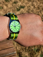 The P-A-R-T-Y? SJT Original wristwatch with a bright green dial and a green-and-yellow striped nylon strap, worn on a person's wrist outdoors with dry grass in the background.