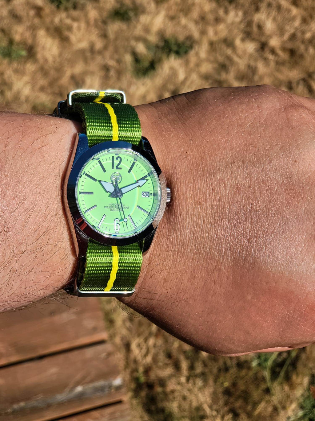 The P-A-R-T-Y? SJT Original wristwatch with a bright green dial and a green-and-yellow striped nylon strap, worn on a person's wrist outdoors with dry grass and a wooden surface in the background.