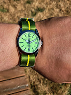 The P-A-R-T-Y? SJT Original wristwatch with a bright green dial and a green-and-yellow striped nylon strap, worn on a person's wrist outdoors with dry grass and a wooden surface in the background.