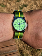 The P-A-R-T-Y? SJT Original wristwatch with a bright green dial and a green-and-yellow striped fabric strap, worn on a person's wrist against an outdoor background of dry grass and a wooden surface.