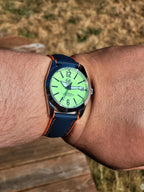 The P-A-R-T-Y? SJT Original wristwatch with a bright green dial and a dark blue textured strap with orange stitching, shown worn on a person's wrist outdoors.