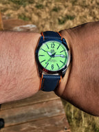 A close-up of a wrist wearing The P-A-R-T-Y? SJT Original wristwatch  outdoors. The watch has a bright neon green dial, a dark blue textured strap with orange stitching, and a dark blue bezel.