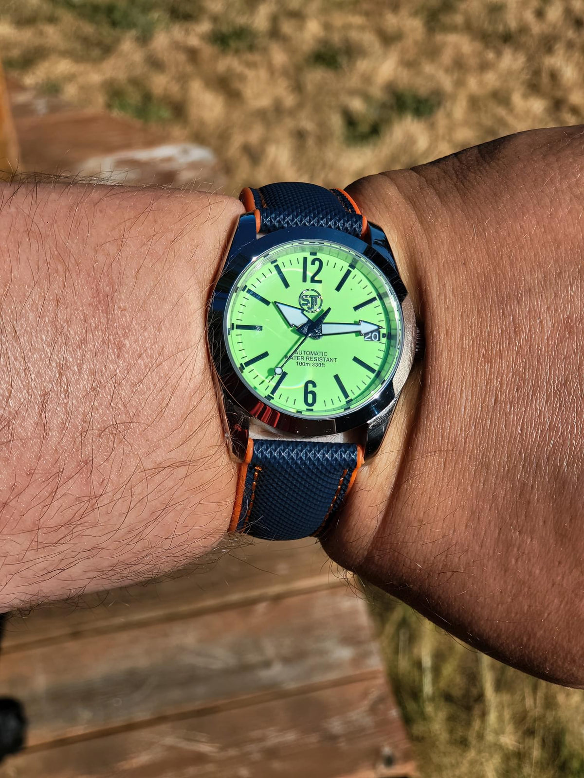 A close-up of a wrist wearing The P-A-R-T-Y? SJT Original wristwatch  outdoors. The watch has a bright neon green dial, a dark blue textured strap with orange stitching, and a dark blue bezel.