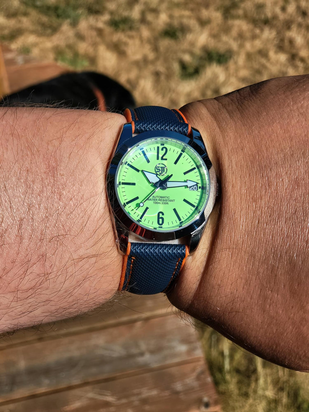 A close-up of a wrist wearing The P-A-R-T-Y? SJT Original wristwatch with a bright green dial, silver-toned case, and dark blue textured strap with orange stitching, photographed outdoors on a wooden surface with dry grass in the background.