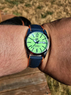 A close-up of a wrist wearing The P-A-R-T-Y? SJT Original wristwatch with a bright green dial, silver-toned case, and dark blue textured strap with orange stitching, photographed outdoors on a wooden surface with dry grass in the background.