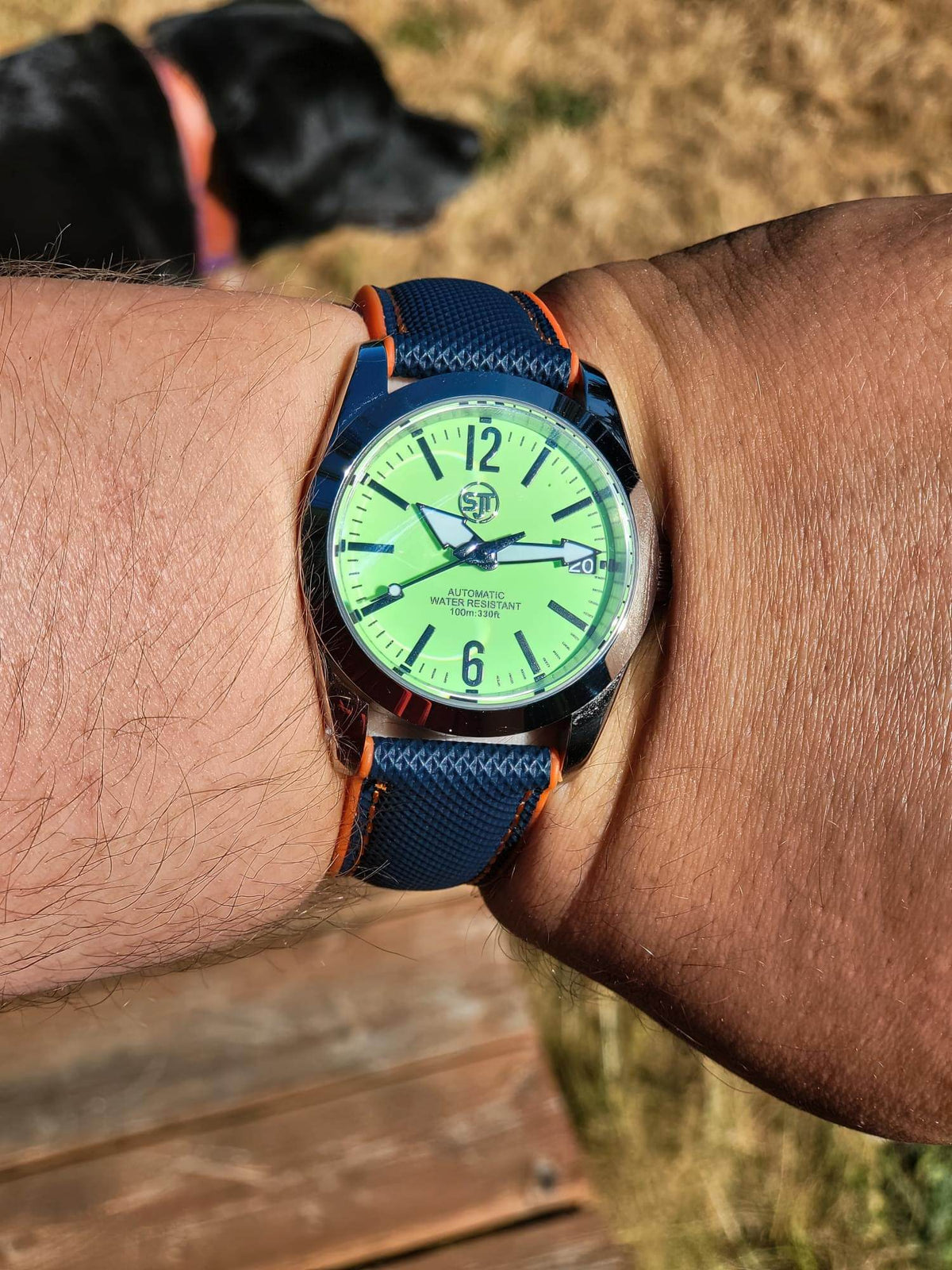 A close-up of a wrist wearing The P-A-R-T-Y? SJT Original wristwatch outdoors. The watch has a bright green dial with black markers and hands, a dark blue textured strap with orange stitching, and a dark blue case.