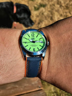 The P-A-R-T-Y? SJT Original wristwatch  with a bright green dial and a blue textured strap with orange stitching, shown worn on a person's wrist outdoors with a dog blurred in the background.