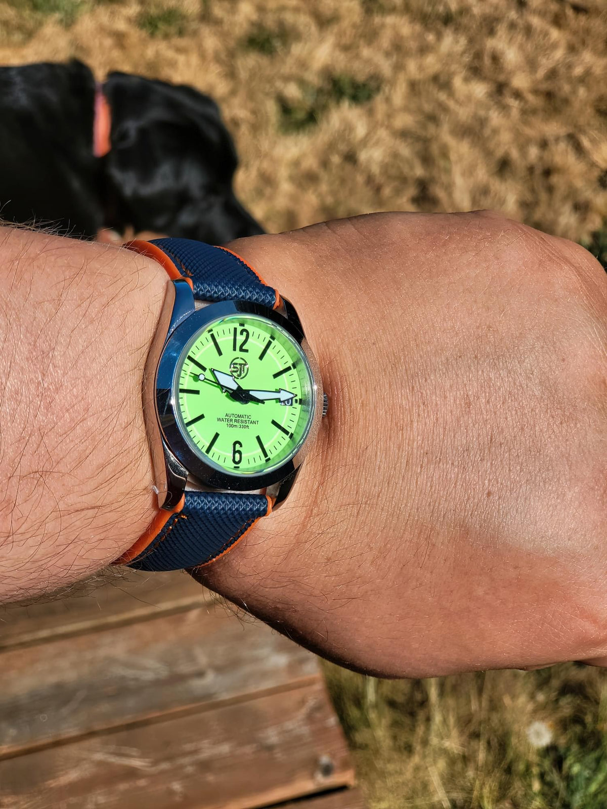 A close-up of a wrist wearing The P-A-R-T-Y? SJT Original wristwatch outdoors. The watch has a bright green dial, a blue textured strap with orange stitching, and a dark blue bezel.