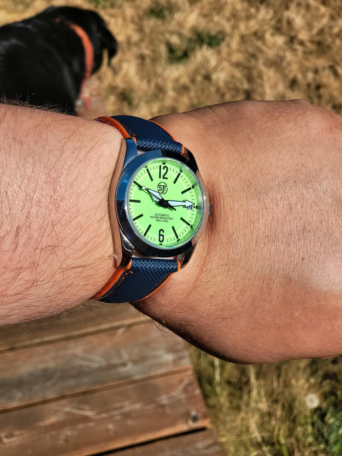 A close-up of a wrist wearing The P-A-R-T-Y? SJT Original wristwatch outdoors. The watch has a bright lime green dial, a blue fabric strap with orange stitching, and a silver-toned case.