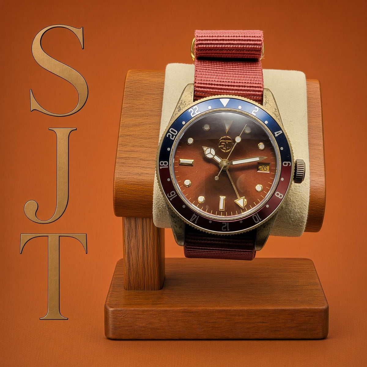 The Vintner SJT Original wristwatch with a brown dial, blue and red bezel, and red fabric strap, displayed on a wooden stand against a warm orange-brown background. Large gold letters 'SJT' appear vertically on the left.