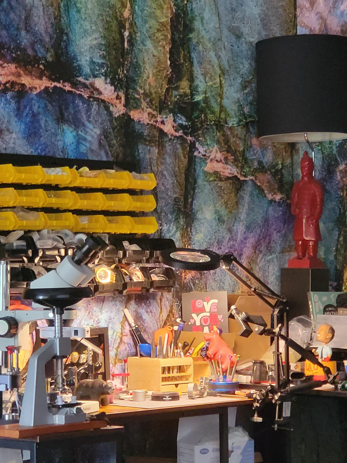 A cluttered workshop or craft station with a large abstract blue and green mural wall behind it. Visible items include a microscope, magnifying tools on an arm, yellow storage bins, a black lamp, a red figurine, and various small tools and containers on a workbench.