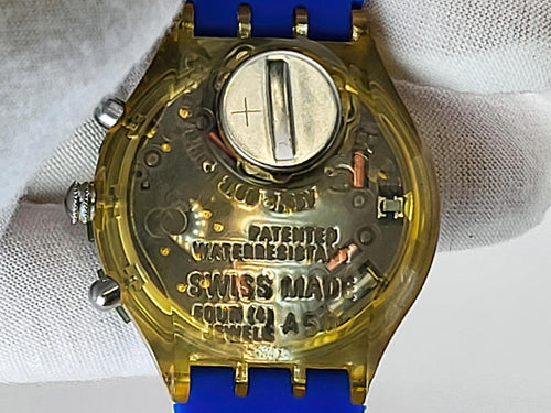 Close-up of the back of a Swatch wristwatch with a transparent gold-toned case, blue strap, and visible internal mechanical details and a battery compartment, resting on white fabric.