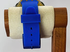 A bright blue silicone Swatch wristwatch strap attached to a gold-tone watch case, displayed on a white felt roll inside a wooden stand against a light gray background.