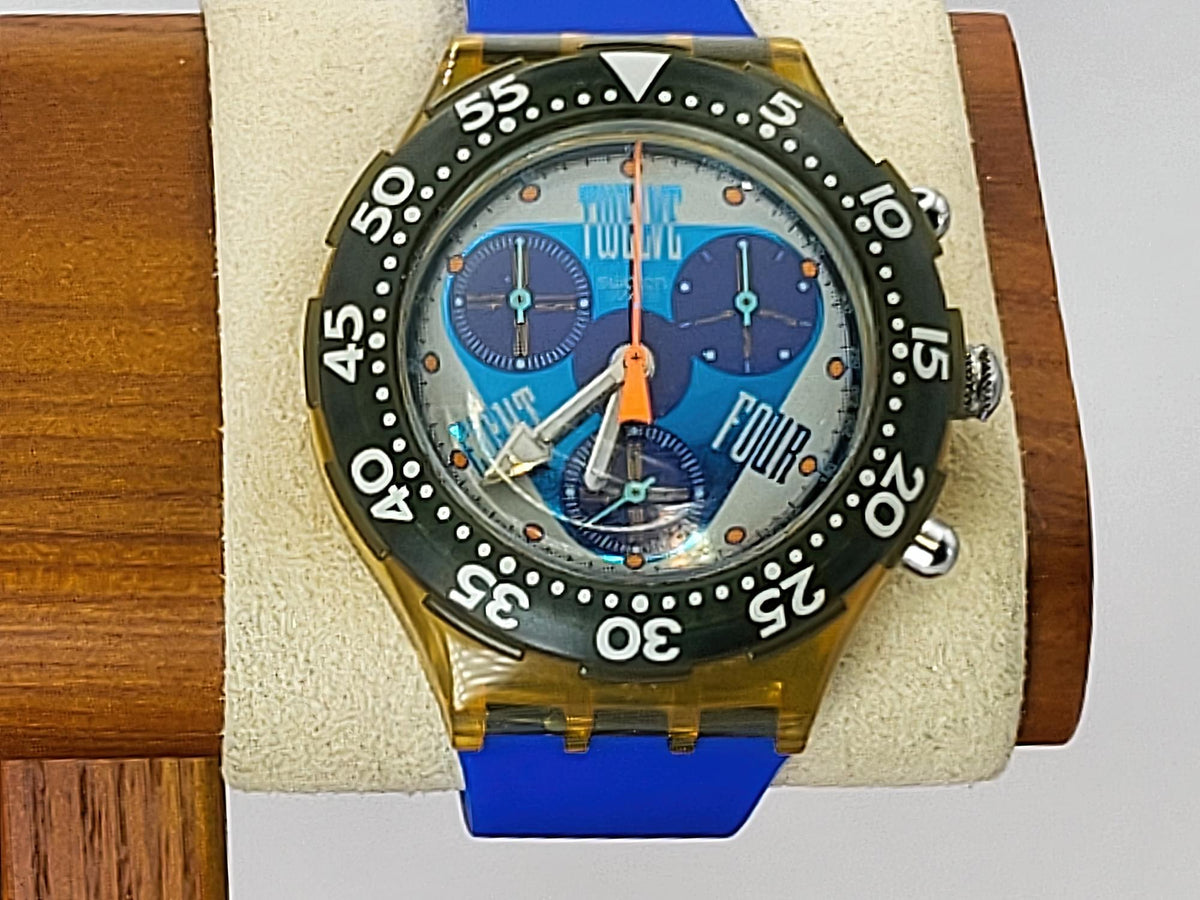 A Swatch wristwatch with a translucent gold-tone case, black bezel, blue strap, and a light blue dial featuring three blue subdials and orange accents, displayed on a cream cushion mounted on a wooden stand.