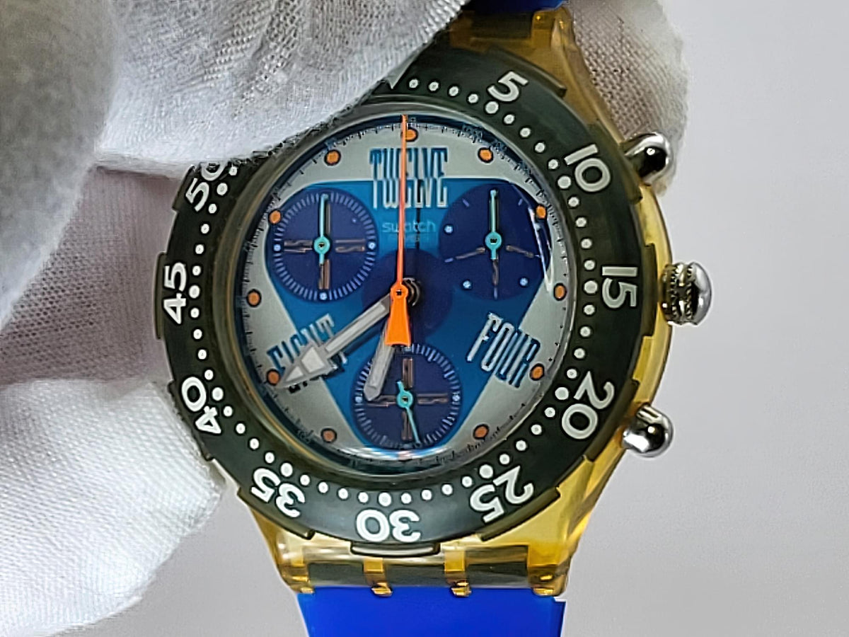 A close-up of a Swatch wristwatch with a translucent gold case, blue and white dial, and blue strap, held by a white gloved hand against a light gray background.