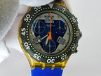 A Swatch wristwatch with a yellow case, a green tachymeter bezel, and a blue dial with orange hands and accents, held by a white fabric sleeve.