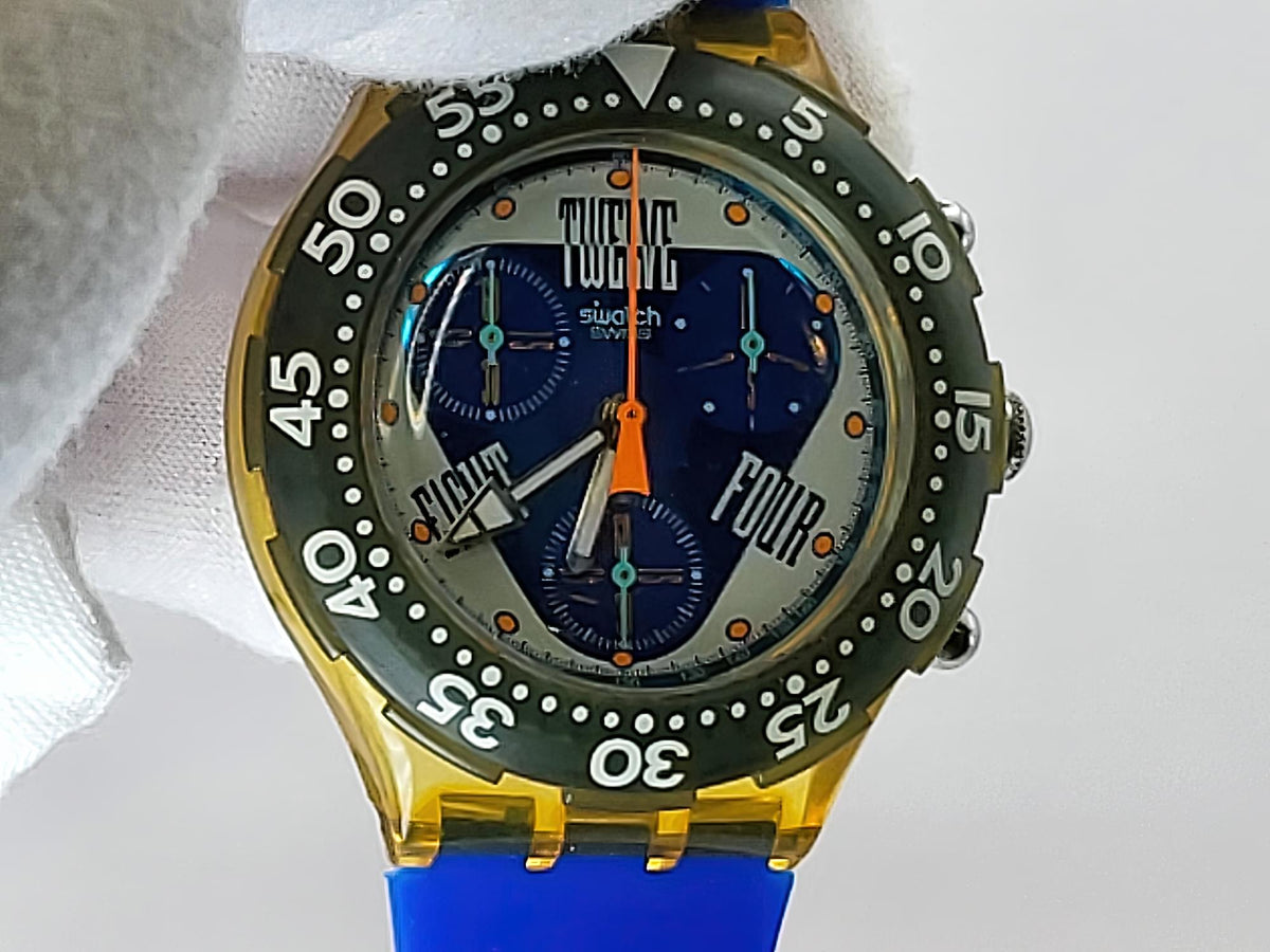 A Swatch wristwatch with a yellow case, a green tachymeter bezel, and a blue dial with orange hands and accents, held by a white fabric sleeve.