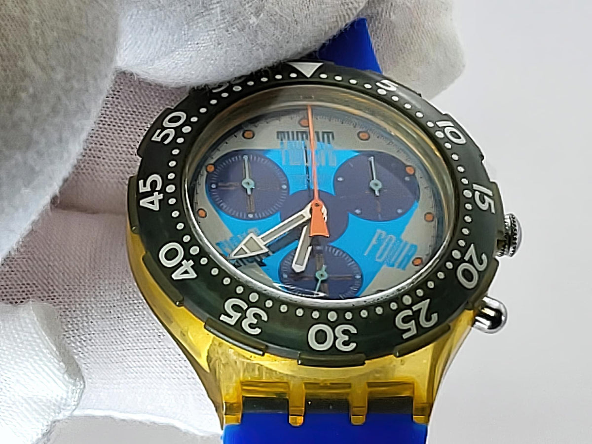 A close-up of a Swatch wristwatch with a translucent yellow case, blue strap, dark green bezel, and a blue dial with three subdials. The watch is being held by a white fabric glove against a light gray background.
